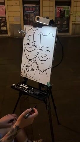 A woman with tattoos on her arm is drawing a caricature on a white paper attached to an easel with a clip. The caricature depicts three figures: two human faces with exaggerated features and a dog's face positioned below them. The woman is holding a black marker and is in the process of drawing the beard of one of the human figures. The easel is lit by a white LED light. The scene appears to be outdoors at night, with a building facade and people walking in the background. The ground is paved. There is a tray attached to the easel containing various pens and markers.