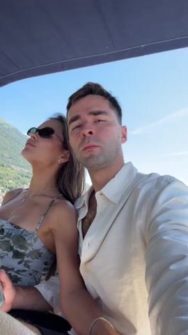 The video shows a close-up, selfie-style shot of a man and a woman sitting next to each other, possibly on a boat given the overhead canopy and the blurred background suggesting water and a hillside. The man, on the right, is wearing a light-colored button-down shirt, partially open, revealing some chest hair. He is looking upwards and to the right with a thoughtful or contemplative expression. The woman, on the left, has her head tilted back, eyes closed, and is wearing sunglasses and a floral print dress. She appears relaxed. Both are looking in the same general direction, upwards towards the sky. The lighting suggests it is daytime, with bright sunlight. The focus is on the couple's faces and upper bodies.