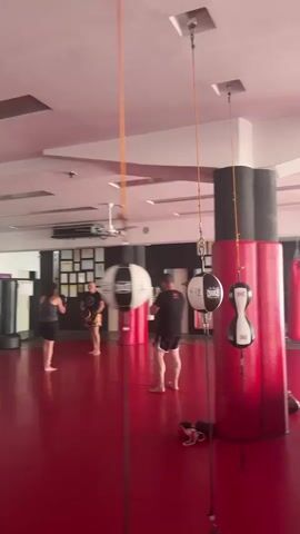 This media file captures an indoor martial arts gym, likely a Muay Thai or boxing facility, in Sant Boi de Llobregat, Spain. The scene is dominated by a vibrant red floor and numerous training apparatus. Suspended from the ceiling are several speed bags, some black and white, others predominantly white with black accents. Adjacent to these, a large, inflatable red and black cylindrical structure stands prominently. In the background, a boxing ring with a metal frame and red padding is visible, along with several heavy punch bags, including red and black varieties, hanging from the ceiling.

Several individuals are present, engaged in training. In the left portion of the frame, a person in black shorts and a black t-shirt is seen barefoot, appearing to be mid-workout. Further back, another individual is visible in similar attire. The lighting suggests it is daytime, and the indoor setting implies the weather is not a factor. The overall atmosphere appears to be one of active training and dedication, with the gym equipment indicating a focus on striking and combat sports. There are no discernible emotions conveyed by the individuals, as they are mostly captured from a distance or their faces are not clearly visible. No text is readily identifiable within the frame, though some branding might be present on the equipment, it is not legible. The presence of multiple training stations and equipment suggests a professional or well-equipped training facility.