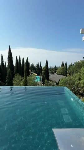 The video shows an infinity pool overlooking a landscape of trees and a clear blue sky. The water in the pool is a vibrant turquoise and ripples gently. In the background, several tall, dark green trees, likely cypress trees, stand out against the lighter green foliage of other trees. Beyond the trees, glimpses of buildings and another, smaller swimming pool with what appear to be water slides can be seen. The sky is a bright, cloudless blue. In the foreground, a person is visible swimming in the infinity pool, their head and shoulders breaking the surface of the water. The camera pans slightly, offering a wider view of the pool and the scenic surroundings. The overall impression is one of a luxurious and relaxing outdoor setting on a sunny day.