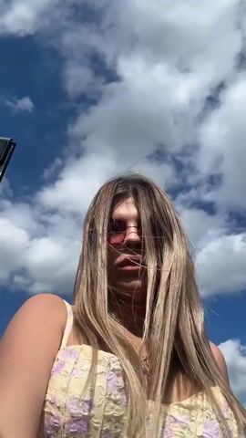 A young woman with blonde hair and pink sunglasses is in the foreground, holding her hair back. She is wearing a yellow top with a floral pattern. The background is a bright blue sky with white clouds. The woman smiles and sticks out her tongue at the camera.