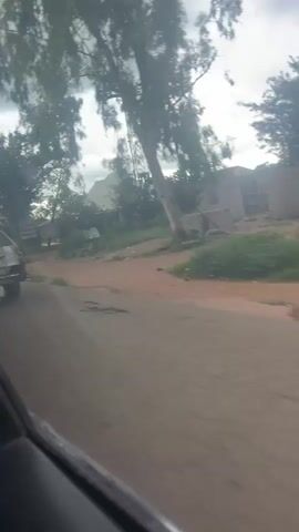 The video shows a view from a moving vehicle. The road appears to be unpaved, with reddish-brown dirt and gravel. On the left side of the road, there is a silver station wagon overloaded with large sacks, tied to the roof rack and extending beyond the vehicle's dimensions. The car appears to be in poor condition, with visible damage to the rear bumper and body. The surrounding environment is rural, with scattered trees and modest buildings. A tall telecommunications tower is visible in the distance. The sky is overcast with clouds.