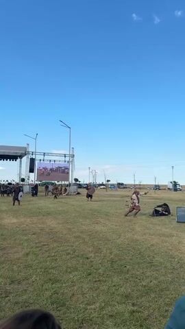 The video shows an outdoor historical reenactment. A group of people dressed in historical costumes, some on horseback, are engaged in what appears to be a staged battle. One person is on a white horse, holding a sword. Another person is on a brown and white horse, wearing elaborate historical armor. There are several other individuals on the ground, some in combat stances, with shields and what appear to be swords. In the background, there is a large stage with a screen, and some vehicles are visible. The setting is a grassy field under a clear blue sky with scattered clouds. The camera is positioned at ground level, focusing on the reenactment with some audience members in the foreground.