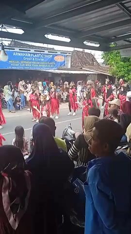 The video shows a parade with dancers in matching red costumes moving on a street. The dancers are mostly young, with headgear that includes large, feathery or bushy decorations. Many people are standing on the sidewalks to watch the parade. They are of various ages and wear different types of clothing. Some are taking pictures or videos with their phones. Behind the crowd is a banner for "ARMAHAM PLASTIC SHOP" with a list of items sold there. The video is shot from under an awning.