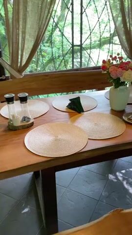 The video shows an outdoor dining area, possibly a restaurant or patio. The camera pans across a long wooden table set with woven placemats, plates, silverware, and vases of flowers. A small cup of coffee sits near one of the settings. The table is positioned in front of a large window overlooking a lush, green outdoor area. The room has a rustic aesthetic, with brick walls and a tiled floor.  The camera continues to pan, revealing a bar area with a wooden counter and shelves, decorated with plants in white pots. The scene concludes with a hanging chair and a view of an outdoor patio with more greenery. The space is well-lit and appears cozy and inviting.