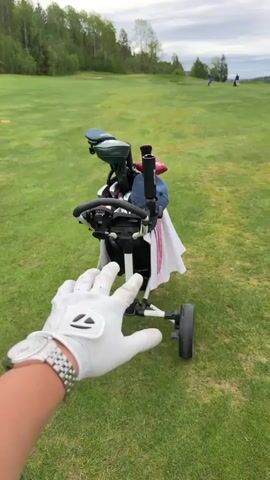 The video shows a person's point of view on a golf course. The person, wearing a white golf glove and a silver wristwatch, reaches towards a white and blue golf cart holding a bag of golf clubs. The cart has three wheels and a handle. The golf bag contains various golf clubs with head covers, a blue baseball cap, and a white striped towel. The golf course is green and grassy, with trees in the background and a cloudy sky. Two people are walking on the course in the distance.