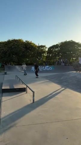 The video captures a skatepark scene at sunset, with a clear, blue sky overhead. Several individuals are engaged in skateboarding within a concrete skatepark, which features ramps and rails. The primary action focuses on two skateboarders performing tricks on a metal rail. One skateboarder, wearing a black t-shirt and black pants, executes a grind on the rail, balancing with arms extended. Another skateboarder, in a light-colored t-shirt and light-colored pants, approaches the same rail. In the background, a group of onlookers stands near a graffiti-adorned wall. Trees line the perimeter of the skatepark, and the warm, low-angled sunlight casts long shadows across the concrete surface, indicating it is late afternoon or early evening. The atmosphere appears relaxed and recreational. No visible text is present within the frame.