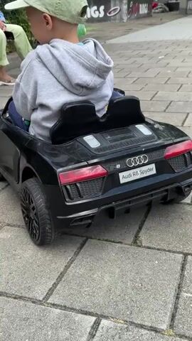 A child wearing a light green baseball cap and a grey hooded sweatshirt is sitting in the driver's seat of a black toy Audi R8 Spyder. The car is parked on a grey cobblestone patio. In the background, an adult wearing a blue striped shirt and green pants is sitting on a wooden bench. The adult is pointing off-camera. The child is holding the steering wheel and looking forward. The car is detailed with red taillights and the Audi logo. The scene appears to be outdoors, with greenery visible behind the bench.