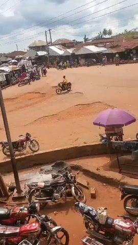 The video shows a bustling marketplace in what appears to be a developing country. The ground is mostly red dirt, and numerous motorcycles are parked in the foreground. In the distance, people move about the market, and various vehicles, including motorcycles, cars, and a minivan, navigate the unpaved roads.  Stalls with umbrellas provide shade for vendors selling goods. The overall atmosphere is active and lively.