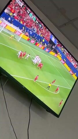 This media file captures a moment of excitement during a football match being broadcast on a television screen. The screen displays a game between Portugal (POR) and Uzbekistan (UZB), with the score currently 1-0 in favor of Portugal at 16:27. The scene on the field shows a group of players in white uniforms clustered around the goal area, with several players in red uniforms positioned defensively. The crowd in the stadium is visible in the background, mostly dressed in red.

The camera then shifts to a close-up of two players in red uniforms celebrating a goal. One player, with a bright smile and wide-open mouth, is the focal point, expressing joy and exuberance. The other player is partially visible behind him, also appearing happy. The backdrop is a vibrant blue, suggesting a professional stadium setting.

The time on the screen advances to 16:31, and the word "FINAL" is visible at the top right, although the score still indicates 1-0 for Portugal. This suggests the broadcast might be showing a replay or a highlights package rather than the live end of the match.

The television itself is a flat-screen, mounted on a wall. Black cables are visible, suggesting it is connected to power and possibly other devices. The overall setting appears to be indoors, possibly a room in Drentsi, Bulgaria, where a football match is being watched. The emotion conveyed is one of triumph and celebration from the scoring team.