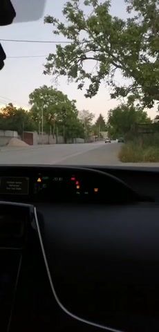 The video shows the interior of a car driving down a paved road lined with trees and houses. The dashboard displays various indicators, including a low fuel warning light and a temperature gauge. The sun appears to be setting, casting a warm glow on the scene. The road is empty except for a few distant cars.