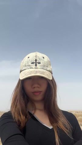 A young woman is featured in a selfie video, smiling gently and looking into the camera. She has long, light brown hair and is wearing a light-colored, possibly denim, baseball cap with a black cross design on the front, a black long-sleeved top with a grey v-neck, and light blue nail polish. The background is a vast, arid, desert-like landscape with sparse, dry, light-colored vegetation and sandy ground. The sky is clear and bright, indicating daytime. As the video progresses, a light-colored, possibly concrete, structure becomes visible in the mid-distance behind her, with faint red flags or banners further in the background. A large, indistinct structure, possibly ancient ruins, can be seen in the far distance, consistent with the location context of Dunhuang City, China. The woman appears calm and pleasant throughout the video.