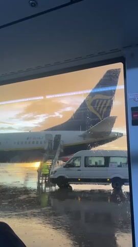 From the vantage point of a well-lit interior, the video shows the exterior of a Ryanair airplane parked on a wet tarmac under a golden sky, possibly at sunset. The plane is white with a dark blue tail adorned with the Ryanair logo. Passengers are embarking or disembarking via stairs attached to the open airplane door. A person wearing a bright yellow vest stands near the base of the stairs. A white van with dark windows and a black number 9752 is parked adjacent to the plane. Rain streaks across the window, and the tarmac glistens with water reflecting the ambient light.