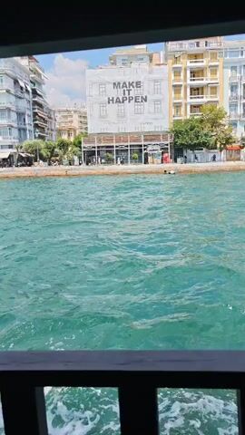 The video captures a view from a moving boat looking towards a waterfront promenade. The water in the foreground is a vibrant turquoise and appears choppy with white foam from the boat's movement. In the background, across the water, is a street lined with multi-story buildings. One prominent building displays a large white banner with the words "MAKE IT HAPPEN" in black lettering. The buildings are a mix of architectural styles, with some featuring balconies and colorful facades. People are visible walking along the promenade and in front of the buildings, and a few cars are parked along the street. The sky is clear and bright, suggesting a sunny day. The overall impression is a scenic view of a coastal city.