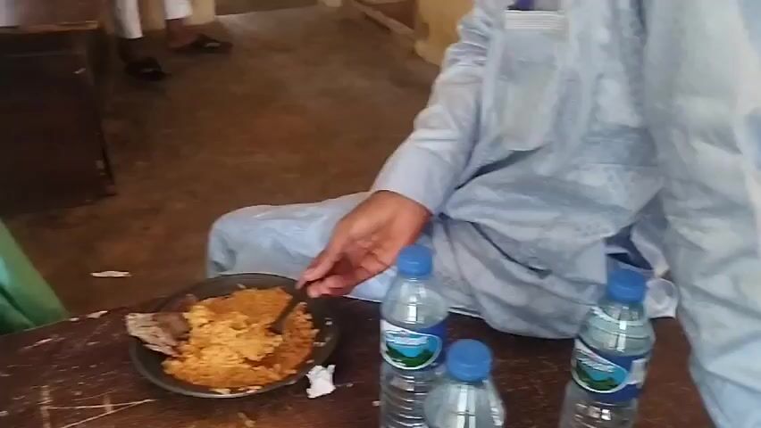 Here is a brief description of the video.

The video shows several men in a room. One man in a light blue outfit is eating something from a tray. Another man in a green outfit and patterned hat looks down, and a man in a white outfit sits in the background. Two water bottles are visible on the table in front of the man in the light blue outfit. The room looks relatively simple and undecorated, with pale-colored walls.