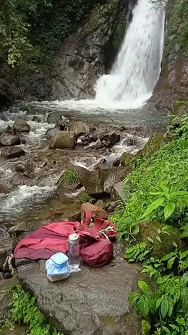 The video shows a natural landscape. A waterfall cascades into a stream filled with rocks. Beside the stream on a large rock, there is a collection of personal items, including a red blanket, a maroon backpack, a clear water bottle, and a blue container. The environment is lush with green vegetation, suggesting a forest or jungle setting. The water flows rapidly over the rocks in the stream.