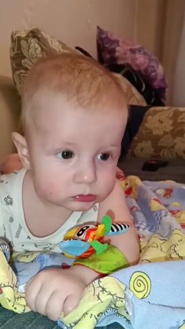 A fair-skinned baby with light brown hair, wearing a white tank top with a pattern, lies on their stomach on a patterned blue blanket, propped up on their elbows. The baby has a colorful stuffed toy on their right arm. In the background, there are pillows with floral patterns. A hand enters the frame and touches the baby's toy. The baby looks at the hand and the toy, smiles briefly, and then looks down.