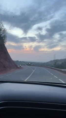 The video shows a view from inside a car driving on a winding mountain road at dusk or dawn. The sky is cloudy with hints of orange from the setting or rising sun. Along the side of the road, at a bend, there are several motorcycles parked and a police van with lights flashing. People are visible standing near the vehicles. The foreground is dominated by the car's dashboard.