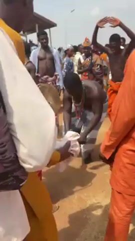 The video shows a group of people engaged in what appears to be a traditional ceremony or celebration. Several men are dancing, some shirtless and wearing cloth wraps around their waists.  One man is playing a drum, and others are clapping and cheering. People are wearing traditional clothing and hats.  The event is taking place outdoors in a dusty area with a structure or building visible in the background. The overall atmosphere seems festive and energetic. The camera moves around, providing different perspectives of the scene.