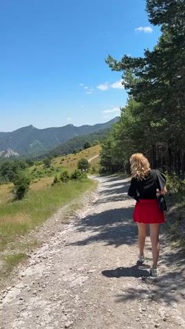 A woman walks down a dirt path in a mountainous, wooded area on a sunny day. She is wearing a black t-shirt, a red skirt, and sneakers. The path is surrounded by green grass, trees, and rolling hills. The sky is a clear blue.