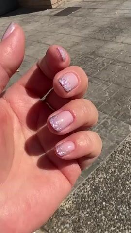 A left hand with a light pink nail polish and iridescent glitter accents on each fingernail is held up in front of a paved surface. The hand is partially illuminated by sunlight, casting shadows.