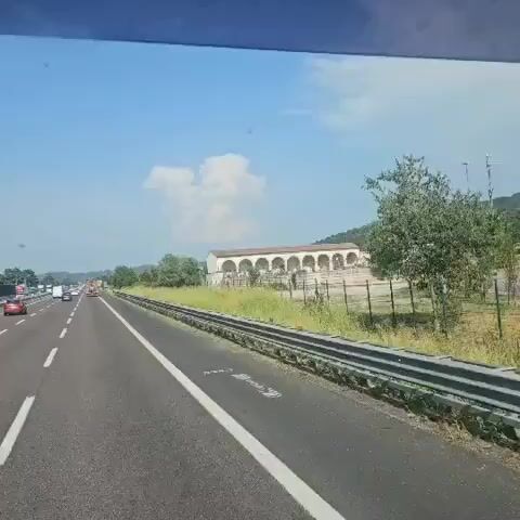 The video shows a view from inside a vehicle traveling on a highway. The road ahead is visible, with cars in the distance. To the right of the highway, there is a building with arched openings, surrounded by a fence and overgrown grass. Trees and a hillside are visible behind the building. The sky is blue with some white clouds. The vehicle appears to be moving at a moderate speed. The overall impression is that of a scenic drive on a sunny day.