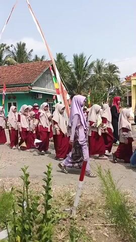A group of children wearing maroon and beige uniforms and headscarves are walking in a procession along a dirt road. Some children are carrying small flags or banners. They are walking past houses and palm trees. A few adults are also present, some of whom appear to be chaperoning the children. There are Indonesian flags displayed on poles. The sky is visible and appears to be bright and sunny.