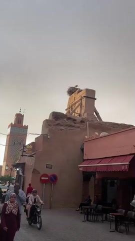 The video shows a large bird, likely a stork, standing in a large nest on top of a dilapidated building. The building is made of a light brown material and appears old, with broken sections and wooden supports holding it up. There are other buildings visible in the background, one with a red awning and another with a green tower. In the foreground, people are walking on the street. The sky is overcast.