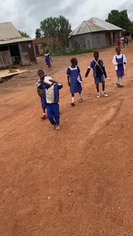 A group of school children, primarily wearing blue and white uniforms, walk along an unpaved red dirt path in what appears to be a rural or semi-rural setting. The sky is overcast, suggesting a cloudy daytime.

In the foreground, several children are visible. One child on the left wears a blue jacket and trousers, carrying a pink bag and holding a book or notebook. Another child in a blue dress with a white collar and cuffs walks with papers in hand. A boy in a blue jacket, dark shirt, and blue shorts, carrying a satchel, walks behind. Further right, a child in a white shirt and blue shorts or trousers walks away from the camera. Other children in similar uniforms are scattered throughout the frame, some looking towards the camera.

The background features simple, one-story buildings with corrugated iron roofs, characteristic of local architecture. A large, prominent cactus-like tree and other sparse vegetation are also visible. The ground is entirely red earth, indicating unpaved roads and paths. The children appear to be casually making their way, possibly to or from school, in a community in Kuru, Nigeria. No discernible text is visible on any objects.