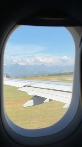 The video shows an airplane wing and a portion of the runway viewed through an airplane window. The wing is white with a pink tip that has "wizzair.com" written on it in white letters. The runway is visible beyond the wing, and there is green grass on either side. In the background, there are mountains under a blue sky with white clouds. The airplane appears to be either on the ground at an airport or just beginning its takeoff or landing sequence. The video is short, about 4 seconds long, and does not contain any people or dialogue.