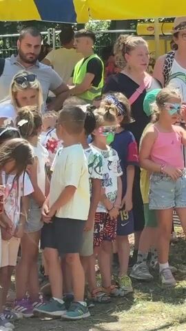 The video shows a group of children and adults gathered outdoors in a park-like setting with trees and grass. Several children in the foreground are wearing colorful masks over their eyes. In the background, a larger group of children and adults are assembled, some of whom are holding what appears to be a rope. The scene suggests an outdoor event or activity, possibly a game or competition. The lighting indicates it is daytime, likely sunny.