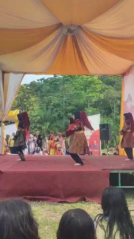 The video shows an outdoor cultural performance. Three dancers in traditional attire, featuring dark headscarves and ornate gold and maroon garments, are performing a coordinated dance on a raised red stage. The stage is covered by a yellow and white tent. The background reveals lush green trees and foliage. An audience, some seated and some standing, watches the performance. In the foreground, closer to the camera, children are present. One child, wearing a brown and orange shirt, is interacting with a white-covered table to the left of the stage. Other audience members are visible behind this child. The overall atmosphere appears festive and communal.