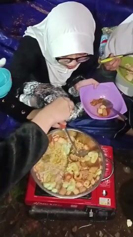 The video shows three people eating a meal. Two of the individuals are seated closely and wearing white headscarves. One person wears glasses. The other person is smiling and eating with a fork and knife from a green plate. The third person’s hand is visible as they stir a large pot of food on a red portable stove. The pot contains a variety of items, including noodles, sliced sausages, and other ingredients. The person in the white headscarf is holding a purple bowl with food and a spoon. A white plate with a design and a spoon is visible nearby. There is a blue tarp beneath them.