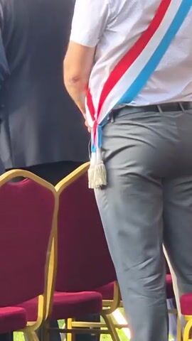 The video shows a group of people standing on a stage outdoors. A man in a white shirt and grey pants, wearing a red, white, and blue sash, stands facing away from the camera. He is on the right side of the stage, with other people to his left. A woman in a blue and white polka dot dress stands on the far left of the stage. The stage is set up on a grassy area, with red chairs arranged on the stage in front of some of the people. There is a white tent structure behind the stage, and trees in the background. Some of the people on stage appear to be speaking or singing, with microphones present. The overall impression is of an outdoor event or ceremony.