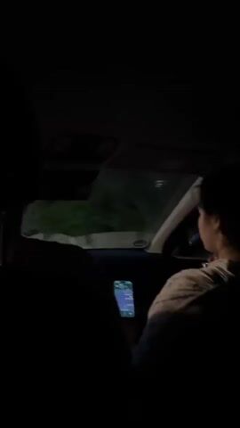 The video shows the interior of a car at night. The camera is positioned behind the front passenger seat, looking forward. In the foreground, the dark silhouettes of two people are visible, one in the driver's seat and one in the passenger seat. The passenger is holding a smartphone, which is illuminated and displaying a screen. Outside the car, through the windshield and side windows, trees and vegetation are faintly visible, dimly lit by external light sources. The overall impression is of a nighttime drive through a wooded area.