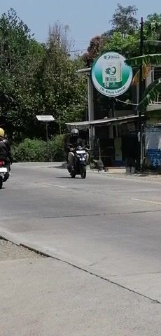 The video captures a street scene with a road on the right side of the frame and trees and buildings on the left. A motorcycle is seen moving from the left to the right. On the left side of the road, there is a circular green sign with text and a logo. Behind the sign, there are some structures that appear to be shops or stalls. A person is riding the motorcycle, wearing a helmet and dark clothing. The environment appears to be outdoors, possibly in a rural or semi-urban area, with a clear sky visible. The image is oriented vertically, with the bottom of the scene appearing on the right.