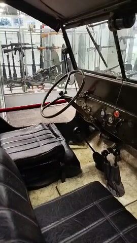 The video pans across the interior of a vintage vehicle, likely a jeep or military truck, displayed in a museum. The focus is on the driver's side, revealing a worn leather seat, a dashboard with gauges and levers, and a steering wheel. In the background, through the windshield, several display cases are visible, filled with an assortment of firearms, suggesting a military history exhibition. The overall environment appears to be well-lit, with a sense of historical preservation.