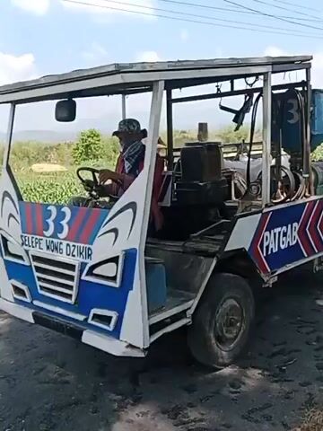 A side view captures a modified vehicle, possibly a small truck or utility vehicle, driving along a paved road. The vehicle has a weathered, utilitarian appearance with a metal frame supporting a canvas roof over the cab area. A man wearing a hat, red shirt, and a face mask is seated in the driver's seat, operating the vehicle. The rear of the vehicle is a flatbed with a metal railing, carrying several large white sacks, likely containing goods. The side of the vehicle is decorated with red and blue chevron patterns and the word "PATGAB" is visible. The vehicle appears to be a mobile processing unit, with a hopper and machinery mounted on the flatbed. The environment is rural, with fields of crops, trees, and utility lines visible in the background. The weather is sunny with a clear sky. The video seems to be filmed from another moving vehicle, possibly a motorcycle, as a mirror is visible in the foreground. The vehicle is in motion, and the camera follows its movement.