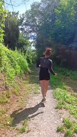 A person with reddish-brown hair tied in a bun is jogging on a dirt path through a wooded area. The person is wearing a dark long-sleeved shirt with a grey design on the upper chest and back, and black shorts. The sun is shining, casting shadows on the path. The path is lined with lush green foliage and trees. The person's arms are extended out to the sides, and their head is turned to the side. The camera is positioned behind and slightly to the side of the person, following their movement.