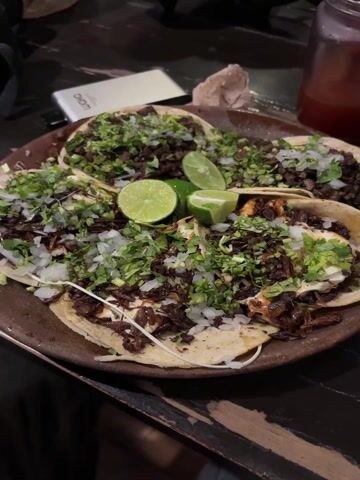 The video captures a close-up shot of a large, round, rustic brown plate laden with several tacos. There are at least five tacos visible, each generously topped with finely chopped fresh cilantro and diced white onions. In the center of the plate, four slices of lime are arranged, consisting of two full slices and two wedges.

The taco fillings vary slightly; the tacos in the back appear to contain dark, diced meat. The tacos in the foreground are topped with a very dark, almost black, textured ingredient, possibly chapulines (grasshoppers) or a type of dark-cooked meat. Beneath these toppings, a white, creamy sauce or melted cheese is visible on some of the front tacos.

The scene is dimly lit, suggesting an indoor setting like a restaurant or dinner table. In the blurred background, a white electronic device (possibly a power bank) with text like "LUOMO" or "LQMO" is visible on the left, alongside a crumpled brown napkin. On the right, a glass containing a reddish-brown beverage is partially visible. The primary focus remains sharp on the plate of tacos.