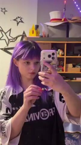 A young person with purple hair and bangs is taking a selfie video in a room. They are holding a phone with a transparent case, and appear to be examining or adjusting a small, colorful object in their other hand. They are wearing a black t-shirt with white writing and a white jacket. Behind them is a white wall decorated with black stars. There is also a wooden shelf filled with various items, including hats and boxes. The room is lit with a soft, slightly purple light, likely from the phone's screen or a nearby light source.