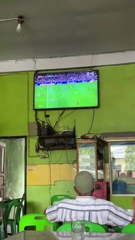A person is seated with their back to the camera, facing a television screen that displays a soccer match. The person is wearing a white and striped shirt and a green cap. The television is mounted on a bright green wall, above a shelf cluttered with wires and electronic equipment. Below the shelf, a small glass cabinet is visible. The environment appears to be a casual establishment with green plastic chairs and tables in the foreground. Other details include a door on the left and a mirror reflecting an outdoor scene on the right.