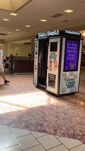 The video shows a man walking in a mall. He is wearing a white t-shirt and shorts. He walks past a photo booth. Then, the camera pans to show a group of people walking down the mall. A Christmas tree is visible in front of a store.