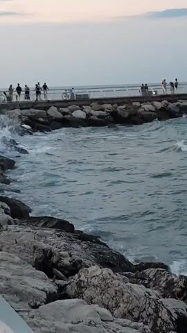 A group of silhouetted figures are visible standing and walking on a pier in the distance. The pier extends over a body of water where waves are crashing against a rocky shore in the foreground. The sky above is a pale, overcast color. The overall scene depicts a coastal environment with people enjoying a view of the ocean.