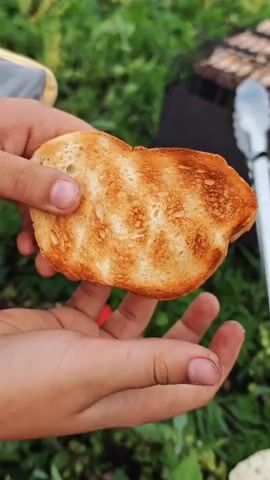 A close-up shot shows a person holding a piece of toasted bread over a grassy area. The bread is golden brown and appears to be slightly torn. In the background, a dark grill and some green foliage are visible.