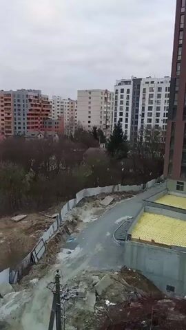 The video shows an urban landscape on an overcast day.  Multiple residential buildings of varying heights and color schemes are visible in the background. In the foreground, a construction site is bordered by a grey metal fence. A narrow, asphalt road curves through the construction area.  A new concrete structure with a yellow, possibly foam, covering is seen next to the road. Scattered piles of dirt, debris, and construction materials are evident. Bare trees and shrubs fill the space between the construction site and the residential buildings. An electrical pole stands to the left of the frame.  The video is taken from an elevated position, likely from within a taller building. No people are visible.