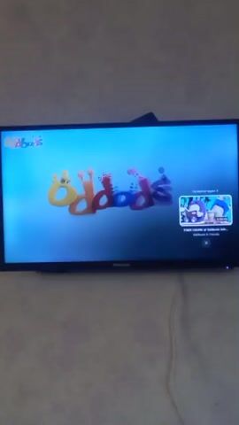 A television screen displays a children's cartoon titled "Oddbods." The show's logo, featuring colorful, bubbly letters, is visible in the top left corner. The main scene shows a purple cartoon character with big eyes and an antenna standing in front of a wall adorned with framed pictures of hands holding paintbrushes. A smaller window in the bottom right corner of the screen shows a thumbnail for another "Oddbods" episode titled "Four Colors of Oddbods."