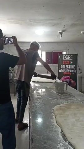 The video shows two men in a brightly lit, possibly industrial kitchen. One man, wearing a black t-shirt and jeans, stands on the left, holding a smartphone in landscape orientation, appearing to record or photograph the scene. The other man, wearing a tan vest over a long-sleeved shirt and a white hairnet, is positioned behind a long stainless steel table. He is bent over the table, using a rolling pin to flatten a large piece of dough. The table is dusted with flour, and there are other pieces of dough visible on the table and to the right of the frame. In the background, a banner with the text "FORF GAZA PALESTINE CHILDREN'S CAKE" is visible, along with what appears to be a pile of colorful flowers. The overall impression is of a baking operation preparing a large cake.