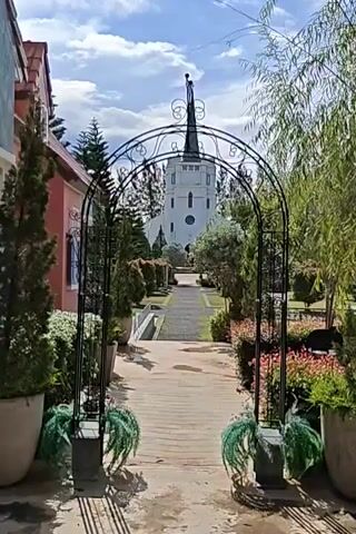 The video shows a pathway leading through an archway towards a white church with a tall, pointed steeple. The pathway is made of bricks and lined with plants and trees. There are potted plants on either side of the archway, and the church is visible in the distance. The sky is blue with some clouds. The overall scene is a well-maintained garden leading to a church.