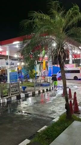 At night, a person in a black shirt and green shorts sweeps the wet ground near palm trees at a gas station. A white van is parked in the background.